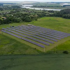 Rozległa działka pod fotowoltaikę z rzędami paneli słonecznych na zielonym polu, w tle wieś i jezioro.