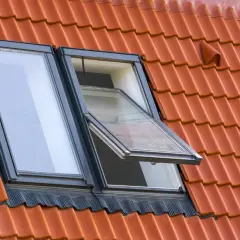 Dwa okna dachowe na czerwonym dachu, jedno otwarte. Standardowe wymiary okien dachowych zapewniają komfort.