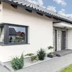 Nowoczesny dom z szarym, narożnym oknem ze słupkiem, kamienną elewacją przy drzwiach i zadbanym ogrodem.