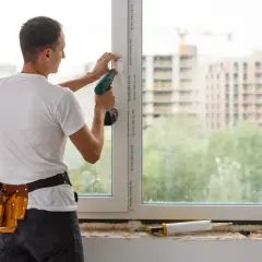 Montażysta przykręca klamkę w nowym oknie. Trwa wymiana okien w bloku, w tle widać budowę.