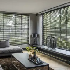 Nowoczesny salon z szarym narożnikiem i stolikiem kawowym. Duże okna z czarnymi żaluzjami wewnętrznymi, które dodają elegancji.