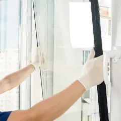 Pracownik w białych rękawiczkach wymienia szybę w oknie. Szyba jest lekko pęknięta, co widać na zdjęciu.