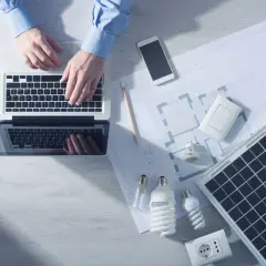 Analiza sprawności paneli fotowoltaicznych na laptopie. Na biurku projekt, żarówki, gniazdko i telefon.