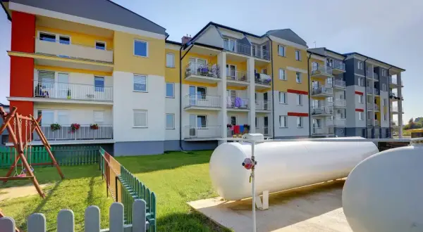 Nowoczesny blok z kolorową elewacją i balkonami. Obok plac zabaw i duże zbiorniki, które zapewniają ogrzewanie gazowe w bloku.
