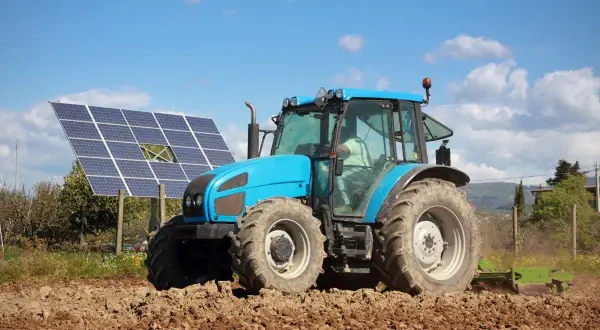 Błękitny traktor orze pole obok paneli fotowoltaicznych. Rolnik korzysta z dofinansowania dla rolników fotowoltaika, inwestując w zieloną energię.