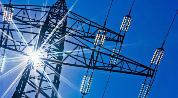 Słońce przebija się przez kratownicę wieży energetycznej, oświetlając izolatory przewodów, które przenoszą prąd dwufazowy.