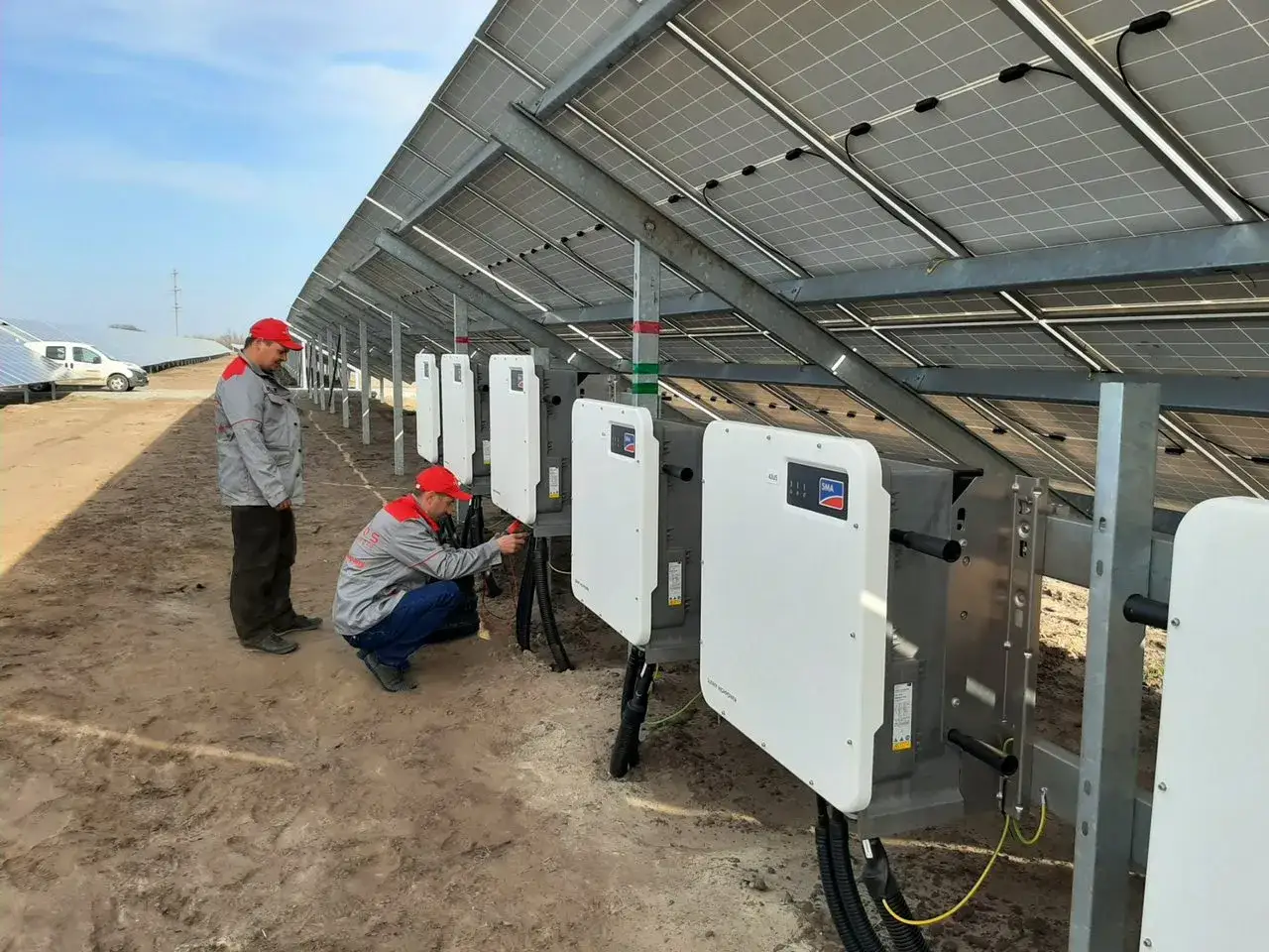 Dwóch techników pracuje przy falownikach na farmie fotowoltaicznej. Instalacja paneli słonecznych w toku.