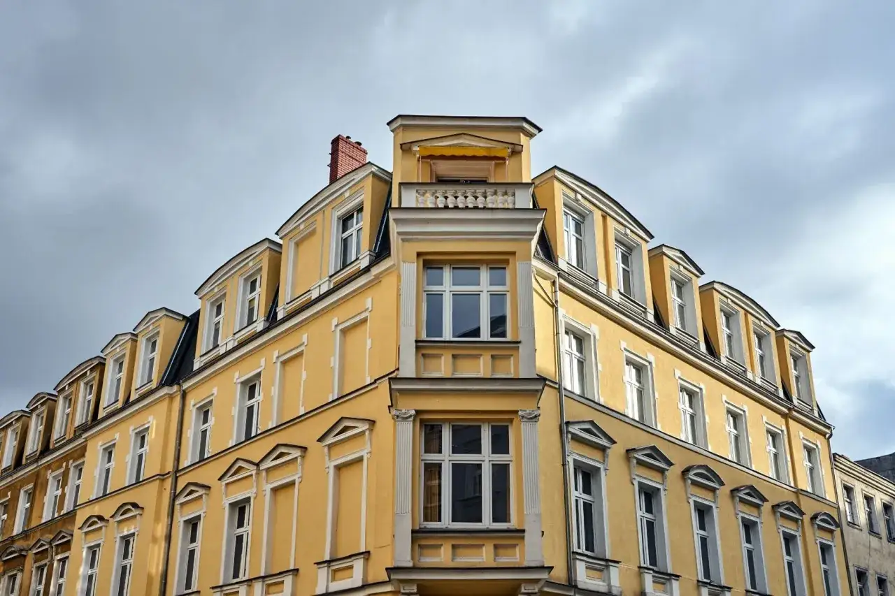 Żółta kamienica z licznymi białymi oknami, w tym ozdobnym wykuszem. Dachówki i komin dodają uroku.