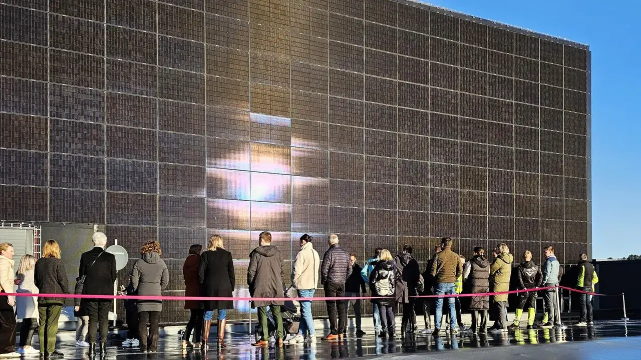 Grupa ludzi ogląda ogromną ścianę pokrytą pionowo ułożonymi panelami fotowoltaicznymi, na której odbija się słońce.